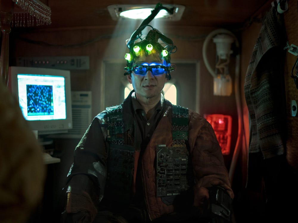 A man wearing a head-mounted device with glowing green lights and blue-tinted glasses sits in a dimly lit, cluttered room with a computer monitor and red emergency lights in the background.