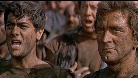 Two rugged, dirt-smeared men with intense expressions stand in the foreground, surrounded by a crowd of similarly distressed individuals in a chaotic, gritty scene.