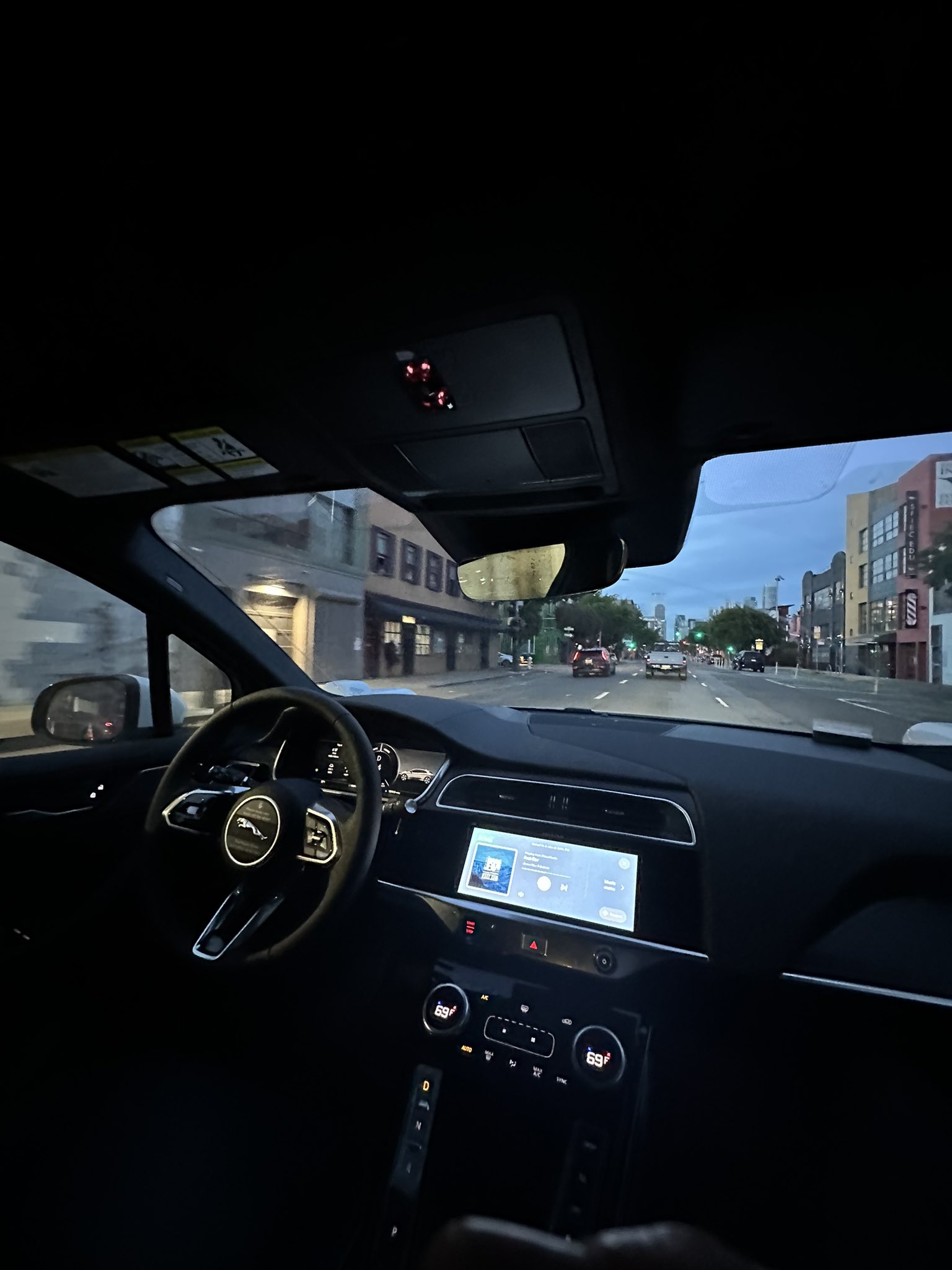 Inside a dark car at dusk, the dashboard glows with a digital display showing 69°F and a music player, while the steering wheel features a Jaguar logo and the view through the windshield reveals a city street with cars and buildings under a twilight sky.