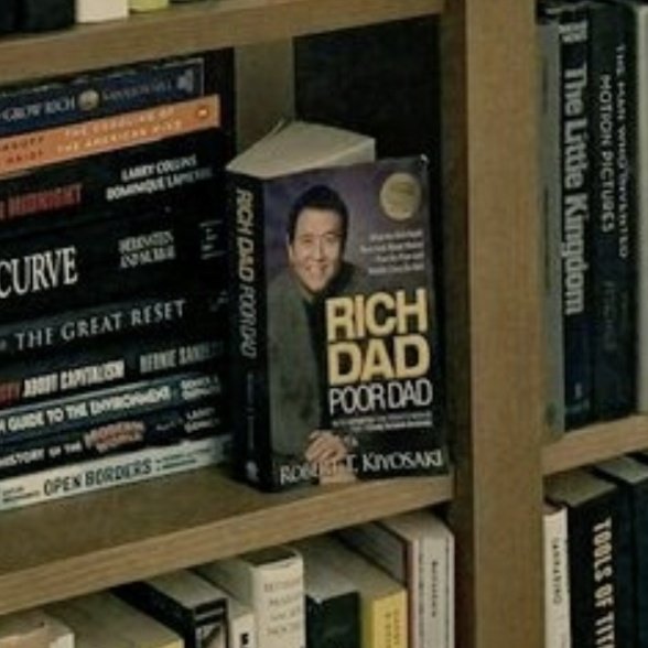 A bookshelf with the book "Rich Dad Poor Dad" by Robert T. Kiyosaki prominently displayed, its cover showing a smiling man, with other books around it including "The Little Kingdom" and "The Great Reset."