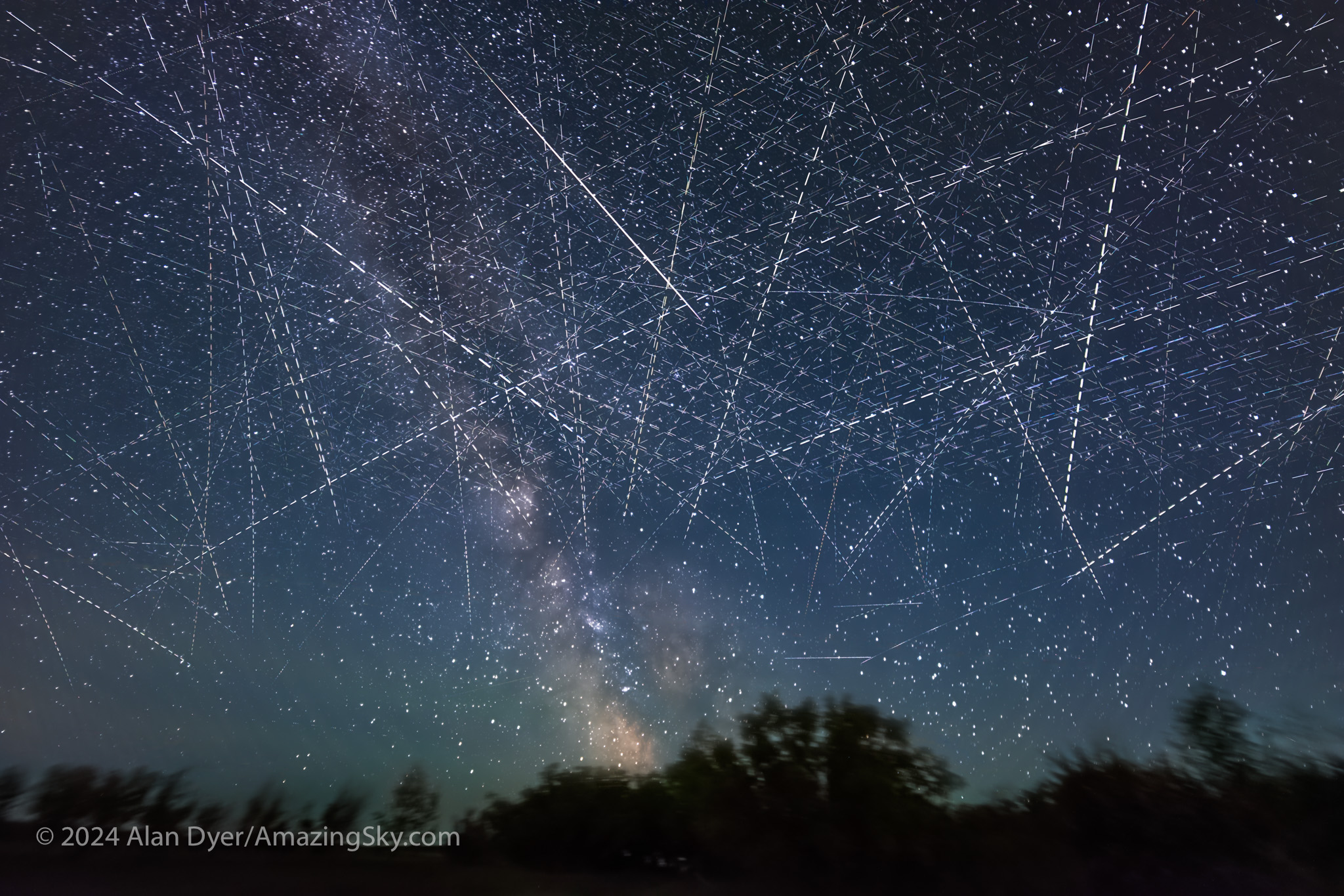 A starry night sky filled with countless twinkling stars and streaks of light from long-exposure photography, with the Milky Way visible in the center and dark silhouettes of trees at the bottom.