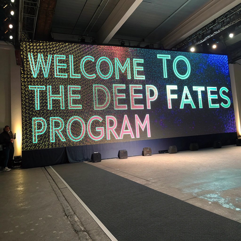 A large LED screen displays "WELCOME TO THE DEEP FATES PROGRAM" in colorful, glowing text against a cosmic background, set in a dimly lit event space with speakers and a runway leading to the stage.