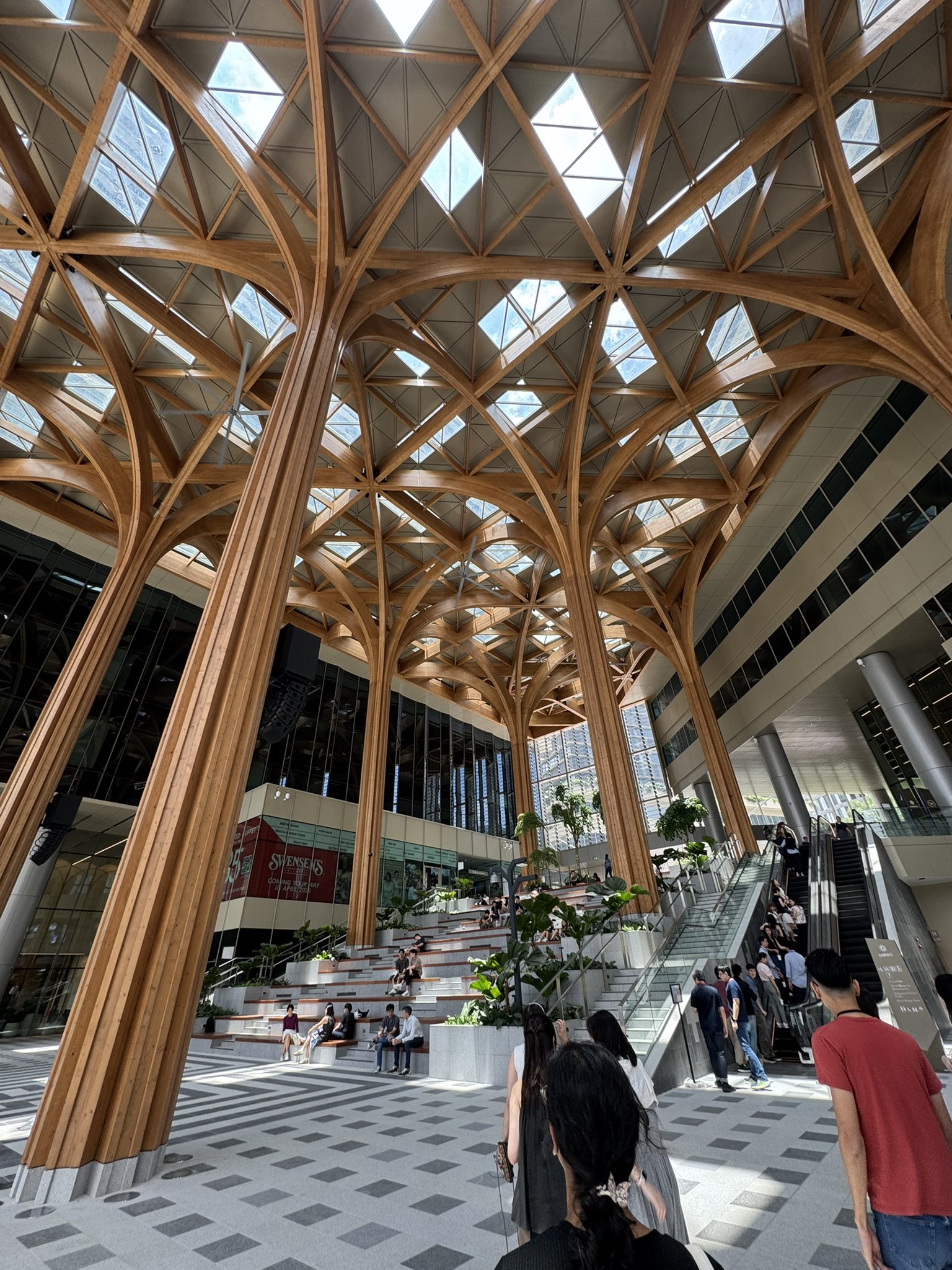 A spacious, modern atrium with a striking wooden ceiling structure featuring intricate beams and skylights, surrounded by people walking and sitting on tiered seating, with glass walls and escalators visible.