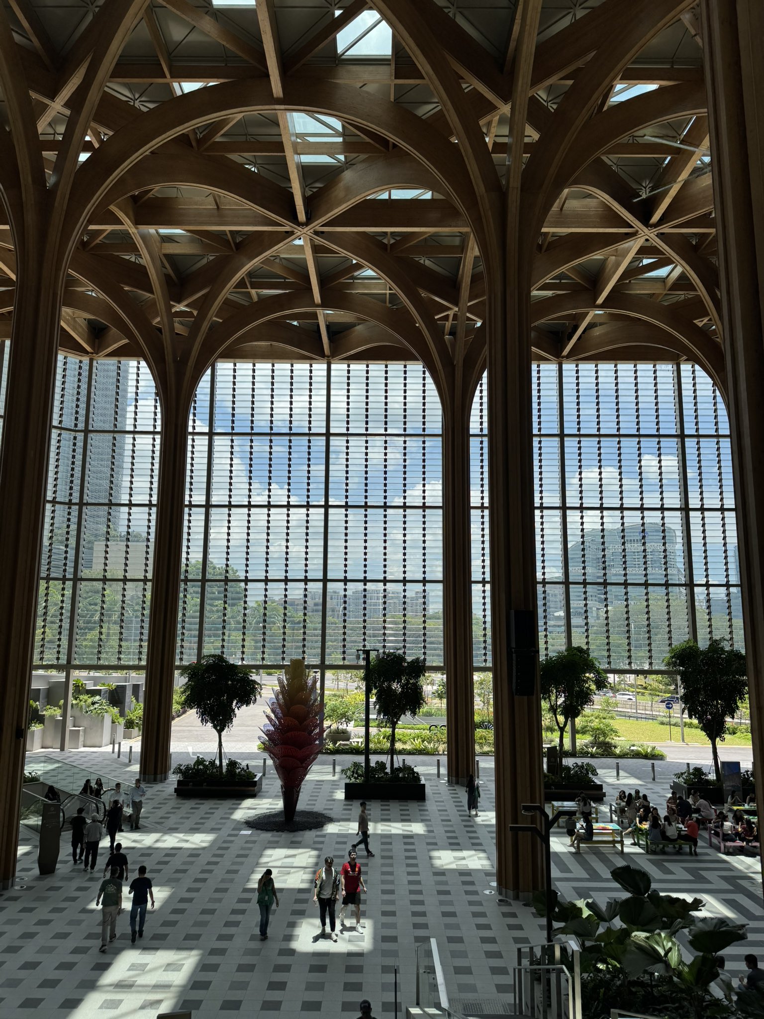 A spacious, modern atrium with a high, arched wooden ceiling and large windows offering views of a city skyline. People stroll and sit on benches beneath a patterned floor, with a central red sculptural tree and potted plants adding vibrant color.