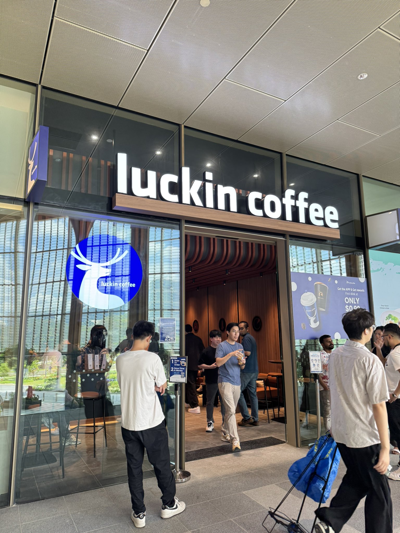 A Luckin Coffee storefront with customers entering and exiting; the glass doors display the brand's logo and a promotional sign for a $0.99 first drink offer.