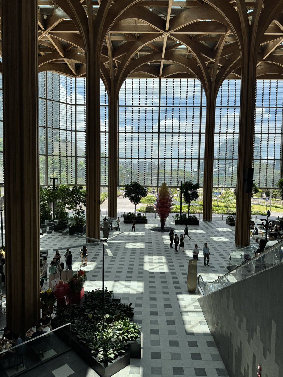 A spacious, modern atrium with a high, arched wooden ceiling and large glass windows overlooking a cityscape, featuring a checkered floor, indoor plants, and people walking or sitting in the sunlit space.