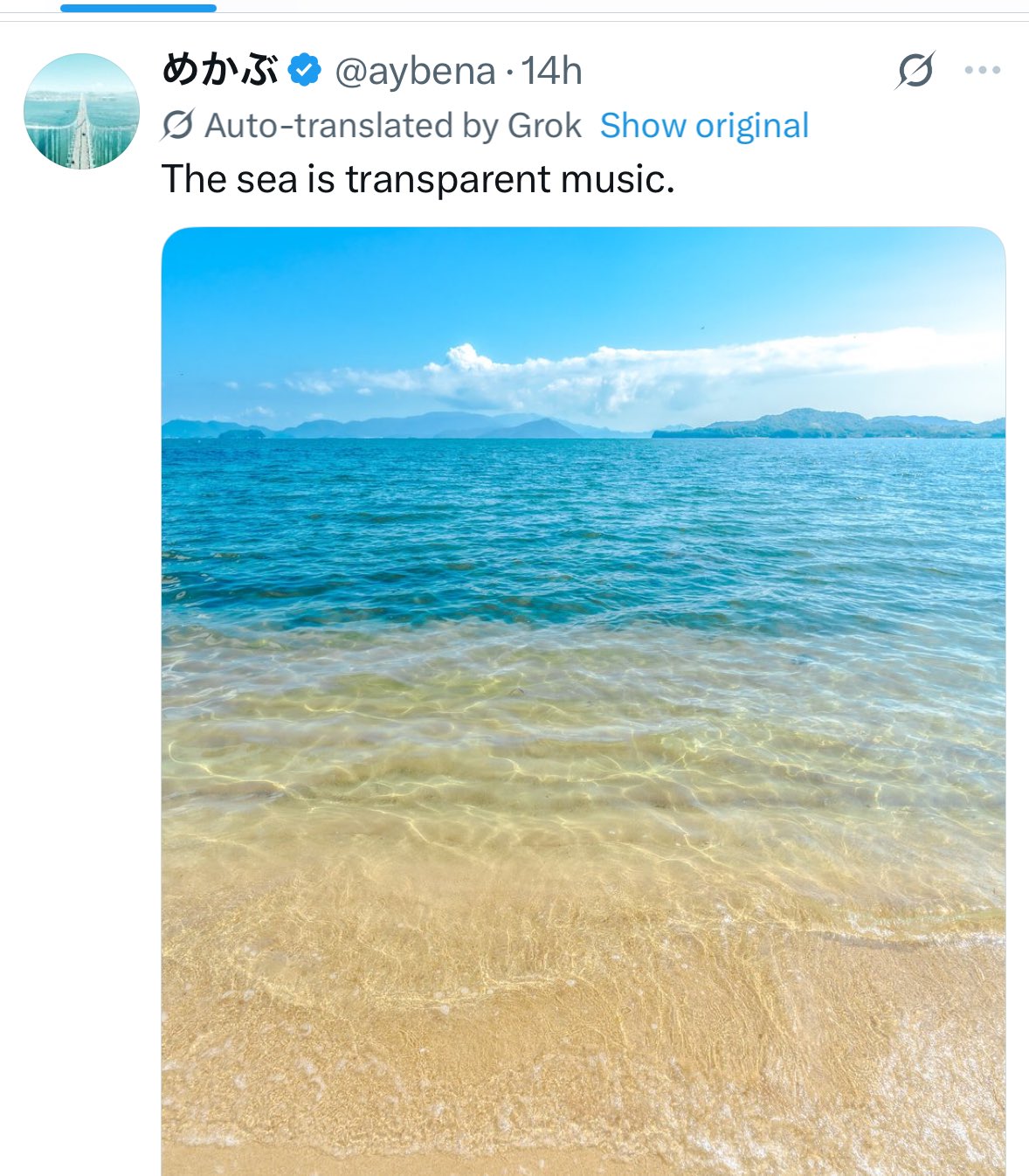 A serene beach scene with clear, shallow turquoise water gently lapping at golden sand under a bright blue sky with scattered clouds. Distant hills and islands are visible on the horizon.