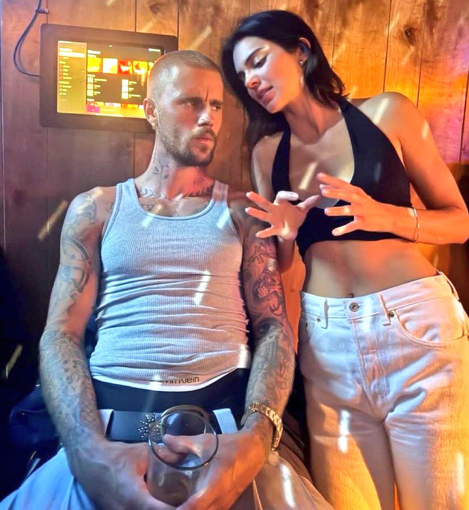 A man with tattoos and a shaved head sits in a dimly lit room, wearing a gray tank top and holding a glass, while a woman in a black crop top and light-colored jeans leans beside him, gesturing with her hands.